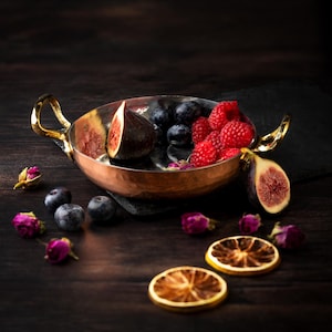 May include: A copper bowl with gold handles, containing figs, blueberries, and raspberries. Dried orange slices and rosebuds are arranged around the bowl on a dark wooden surface, creating a still-life arrangement.