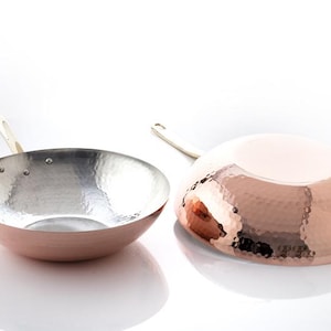 May include: Two copper woks with silver interiors and handles. One wok is upright, showcasing the hammered texture of the interior. The other wok is inverted, displaying the copper exterior.