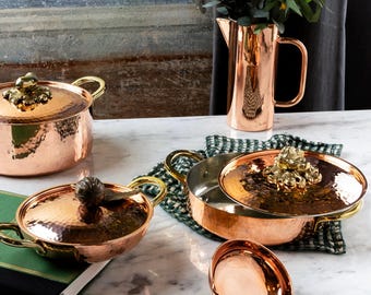 May include: A collection of copper cookware, including pots, pans, and a colander, with brass handles and decorative lid finials. A copper pitcher and wooden-handled ladle are also present. The items are arranged on a marble surface.