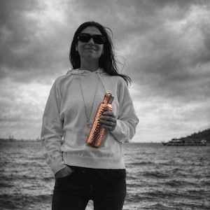 May include: A cylindrical copper water bottle with a textured, hammered finish and a matching metallic cap. The bottle features a distinct metallic sheen that contrasts with the surrounding black and white environment, highlighting its reflective surface.