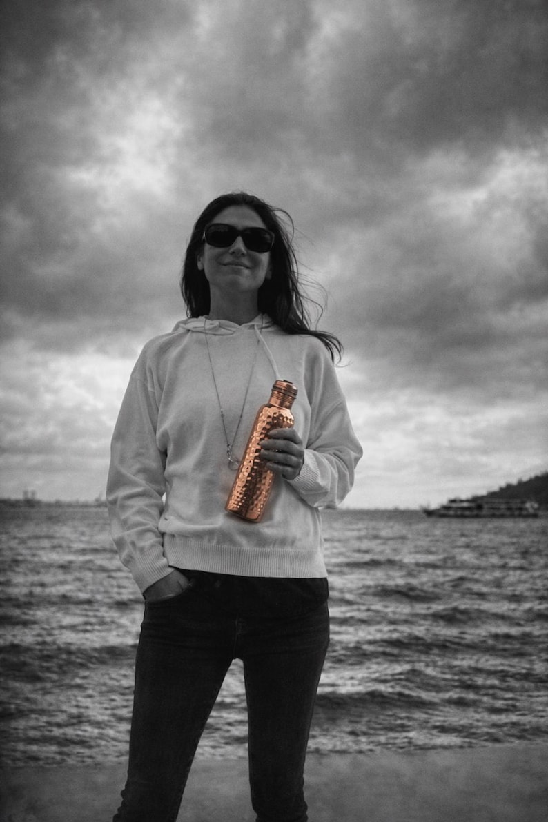 May include: A cylindrical copper water bottle with a textured, hammered finish and a matching metallic cap. The bottle features a distinct metallic sheen that contrasts with the surrounding black and white environment, highlighting its reflective surface.