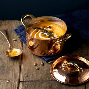Puede incluir: Una olla de cobre llena de sopa naranja, adornada con semillas y un remolino de crema. Una tapa a juego y una cuchara están cerca sobre una superficie de madera. La olla tiene asas de latón.