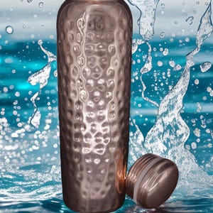 May include: A cylindrical copper water bottle with a textured, hammered finish and a matching screw-top lid. The bottle is positioned upright with its lid resting beside it, surrounded by dynamic splashes of clear water against a blue background.