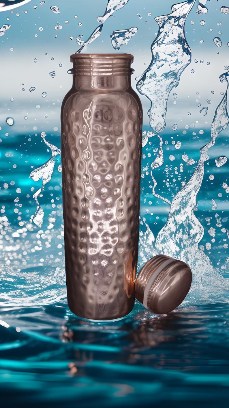 May include: A cylindrical copper water bottle with a textured, hammered finish and a matching screw-top lid. The bottle is positioned upright with its lid resting beside it, surrounded by dynamic splashes of clear water against a blue background.