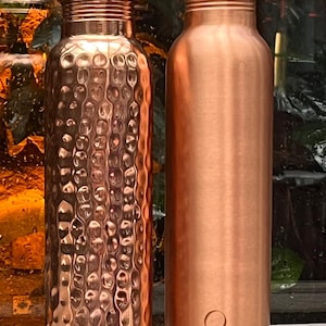 May include: Two copper water bottles standing side by side. The bottle on the left features a textured, hammered finish, while the bottle on the right has a smooth, polished surface with a small letter Q engraved near the base. Both have caps.