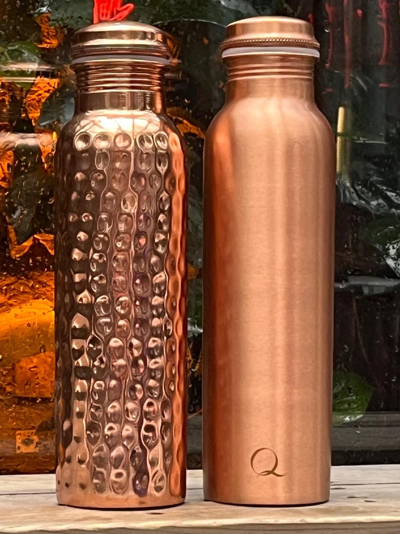 May include: Two copper water bottles standing side by side. The bottle on the left features a textured, hammered finish, while the bottle on the right has a smooth, polished surface with a small letter Q engraved near the base. Both have caps.