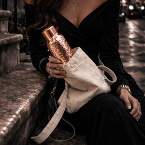 May include: A metallic copper-coloured water bottle with a hammered texture and a smooth cylindrical cap, partially inserted into a light beige canvas drawstring bag featuring two small gold-toned metal buttons near the top opening.