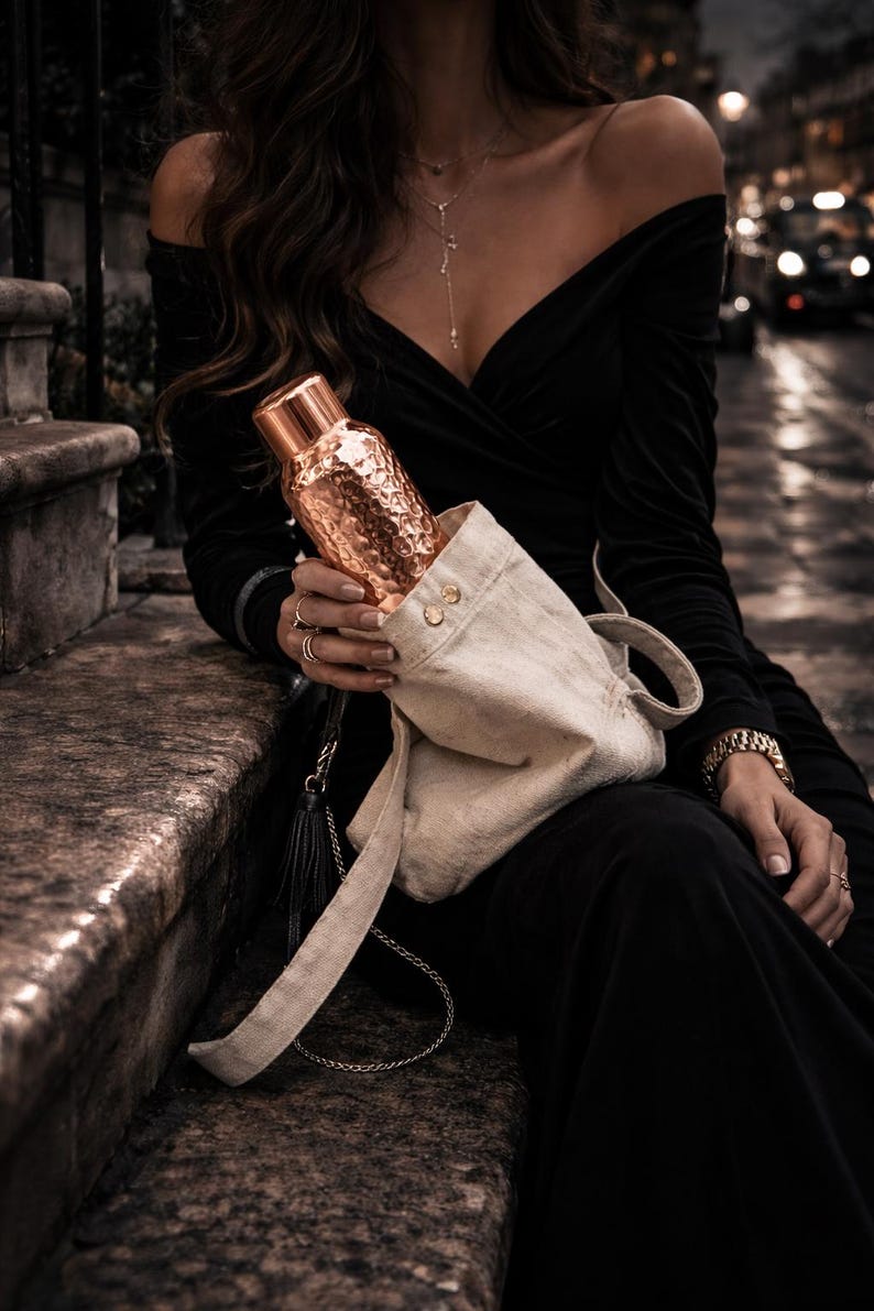 May include: A metallic copper-coloured water bottle with a hammered texture and a smooth cylindrical cap, partially inserted into a light beige canvas drawstring bag featuring two small gold-toned metal buttons near the top opening.
