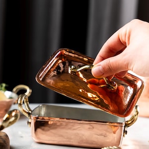 May include: A copper rectangular casserole dish with a lid being lifted. The dish has a hammered texture and gold-colored handles. The interior appears to be silver-colored. The lid has a matching gold handle.