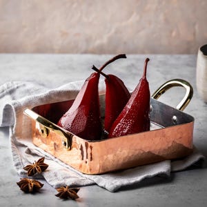 May include: Three red pears poached in a copper baking dish. The pears are arranged in a row with their stems pointing towards the top of the image. The dish is sitting on a white linen cloth.