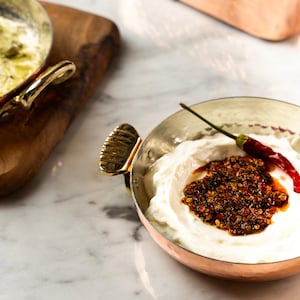 May include: A copper bowl filled with white cream, topped with a dark red-brown sauce and a red chilli pepper. The bowl has a gold handle. Another bowl with a gold handle and a wooden board are in the background.