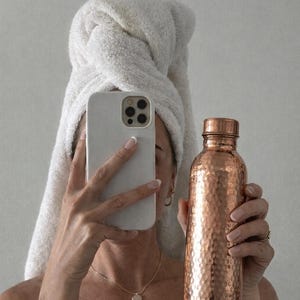 May include: A hammered copper water bottle with a matching screw-top lid. The bottle features a textured, dimpled surface that reflects light, showcasing a metallic reddish-brown finish. The cylindrical container is held upright, displaying its metallic sheen.