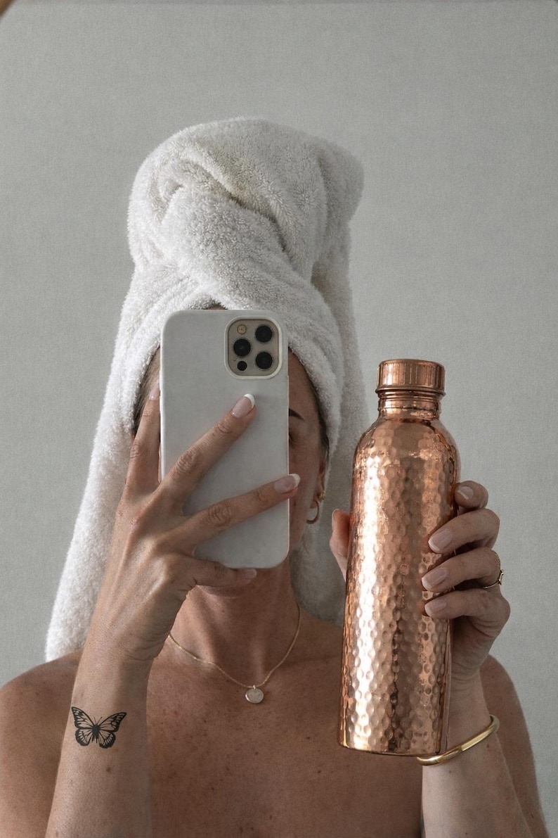 May include: A hammered copper water bottle with a matching screw-top lid. The bottle features a textured, dimpled surface that reflects light, showcasing a metallic reddish-brown finish. The cylindrical container is held upright, displaying its metallic sheen.