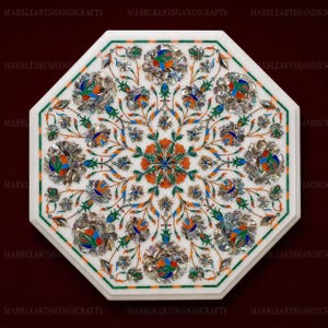Peut inclure: Un plateau de table octogonal en marbre blanc orné d'une incrustation complexe de motifs floraux colorés. La surface présente une fleur centrale orange et bleue entourée de vignes vertes, de petites fleurs bleues et de grands pétales en nacre.