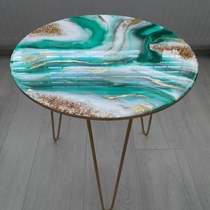 Peut inclure: Une table d'appoint ronde avec un plateau en résine brillante présentant un motif tourbillonnant de vert émeraude, de blanc et d'accents métalliques dorés. La surface est lisse et réfléchissante avec des paillettes dorées. Elle repose sur trois pieds en épingle en métal doré.
