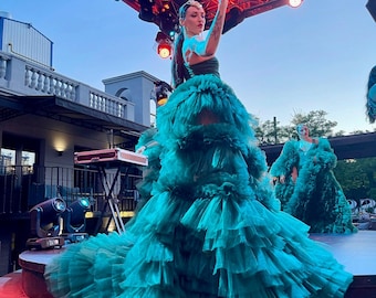 May include: A woman wearing a teal, multi-layered tulle gown with a sparkling crown on her head. She is performing on a stage with red lighting.
