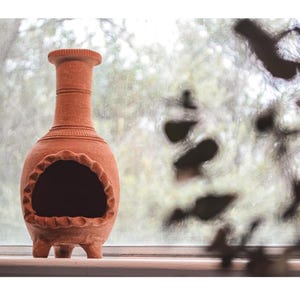 Pu&ograve; includere: Un focolare in terracotta con un'apertura rotonda e tre gambe. Il focolare &egrave; appoggiato su un davanzale della finestra con uno sfondo sfocato.