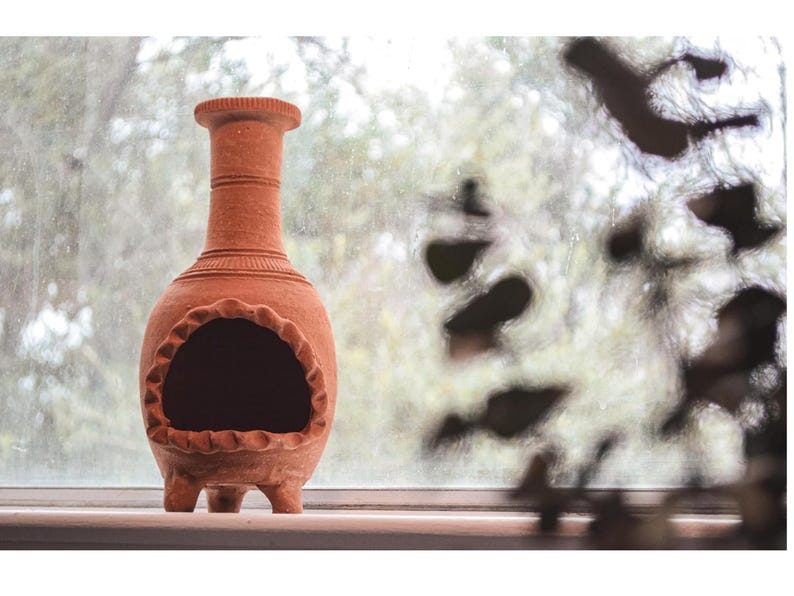 Pu&ograve; includere: Un focolare in terracotta con un'apertura rotonda e tre gambe. Il focolare &egrave; appoggiato su un davanzale della finestra con uno sfondo sfocato.