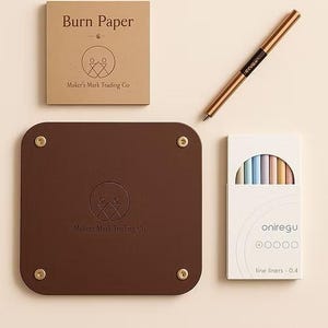 May include: A selection of stationery items, including a brown leather tray with brass studs, a box labelled "Burn Paper", a gold pen, and a box of pastel fine liners. The tray and paper box feature the "Maker's Mark Trading Co" logo.