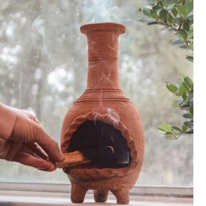 Pu&ograve; includere: Un bruciatore di incenso in terracotta con un'apertura rotonda e quattro gambe. Una mano tiene un bastoncino di incenso sopra l'apertura.