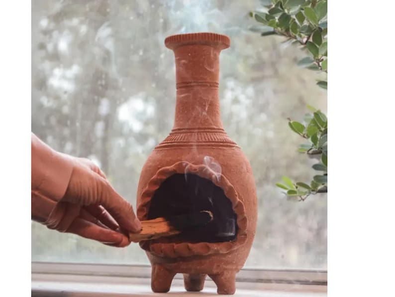 Pu&ograve; includere: Un bruciatore di incenso in terracotta con un'apertura rotonda e quattro gambe. Una mano tiene un bastoncino di incenso sopra l'apertura.