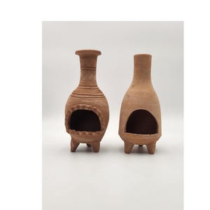 Pu&ograve; includere: Due bruciatori di incenso in terracotta con un'apertura rotonda e tre gambe. I bruciatori sono di colore marrone e hanno un aspetto rustico.
