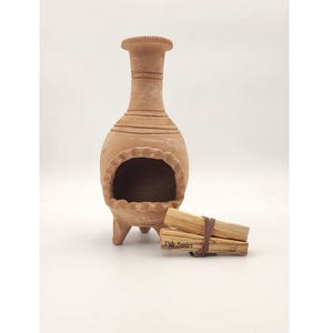 Pu&ograve; includere: Un bruciatore di incenso in terracotta con tre gambe e un'apertura rotonda. Il bruciatore &egrave; fatto di argilla e ha un aspetto rustico e terroso. Due bastoncini di Palo Santo sono davanti al bruciatore.
