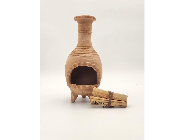 Pu&ograve; includere: Un bruciatore di incenso in terracotta con tre gambe e un'apertura rotonda. Il bruciatore &egrave; fatto di argilla e ha un aspetto rustico e terroso. Due bastoncini di Palo Santo sono davanti al bruciatore.