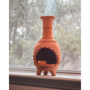 Pu&ograve; includere: Un bruciatore di incenso in terracotta con un'apertura rotonda e tre gambe. Il fumo sale dall'apertura.