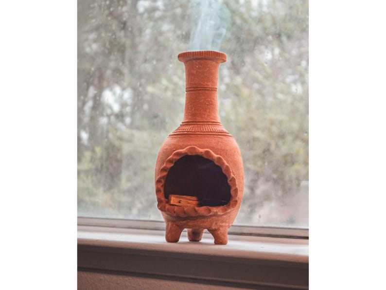 Pu&ograve; includere: Un bruciatore di incenso in terracotta con un'apertura rotonda e tre gambe. Il fumo sale dall'apertura.