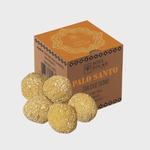 May include: A brown box with gold detailing containing five light-yellow Palo Santo Smudge Bombs. The box is labelled "Soul Sticks" and "Palo Santo Smudge Bomb". The smudge bombs are round and have a textured surface.