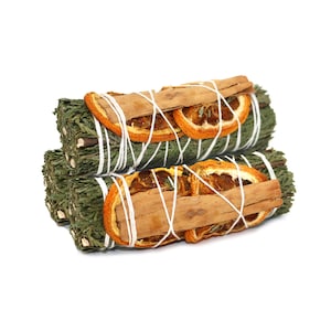Op de afbeelding: Twee bundels groene salie, versierd met gedroogde sinaasappelschijfjes en kaneelstokjes, zijn vastgebonden met wit touw. De saliebundels zijn op elkaar gestapeld, wat een visueel aantrekkelijke opstelling creëert.