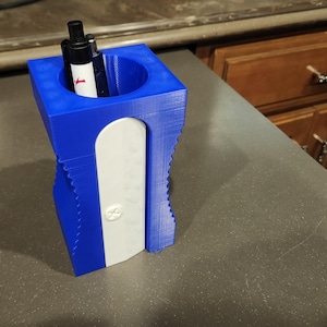 May include: A blue plastic pencil holder with a built-in pencil sharpener. The holder has a round opening at the top for pencils and a white sharpener on the side. A black pen is inserted in the holder.