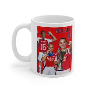 Könnte beinhalten: Weißer Keramikbecher mit rotem Hintergrund und Bildern von Fußballspielerinnen. Der Becher hat einen Griff auf der linken Seite. Die Spielerinnen tragen rot-weiß gestreifte Trikots. Der Becher trägt den Namen "McCabe" und die Nummern "11" und "15".