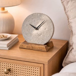 May include: A round, gray concrete clock with black hands sits on a wooden stand. The clock is displayed on a wooden nightstand next to a lamp with a beige lampshade. The nightstand has a woven drawer and a gold knob.