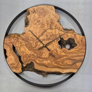 Op de afbeelding: Een ronde wandklok met een natuurlijke olijfhouten wijzerplaat met donkerbruine en bruine tinten. De wijzers zijn zwart en het hout is geplaatst in een zwarte metalen ring. Uniek, rustiek ontwerp.