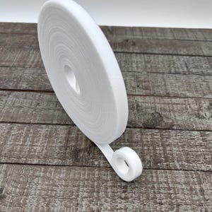 May include: A roll of white hook and loop tape, also known as Velcro, is displayed on a wooden surface. The tape is rolled up, with a small section unrolled, showing the textured surface. The background is a plain white.