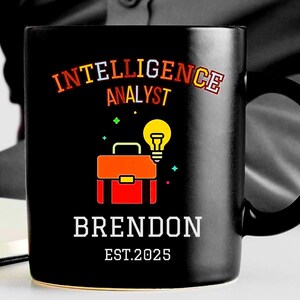 May include: Black coffee mug with the text "Intelligence Analyst" in rainbow colors, a lightbulb, a briefcase, and the name "Brendon" with the text "Est. 2025" below.