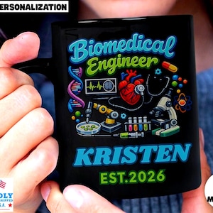 May include: Black ceramic mug with the words "Biomedical Engineer" in blue and green, along with a colorful graphic of medical equipment and a heart. The name "Kristen" and the year "EST.2026" are below. The mug is held by a person.