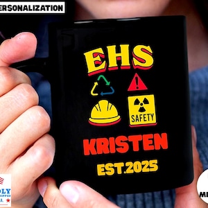 May include: Black ceramic mug with the text "EHS SAFETY KRISTEN EST.2025" in yellow and red. The mug features safety symbols including a recycling symbol, a hazard sign, and a radiation symbol. The mug is held by a person.