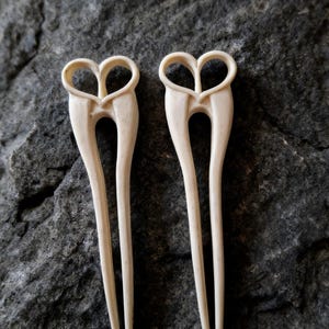 May include: Two ivory-colored hair forks with heart-shaped tops. The hair accessories are displayed against a dark gray, textured background. The forks have a slender, elongated design, suitable for various hairstyles.