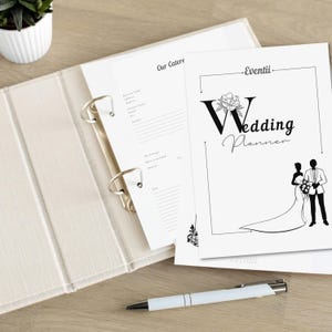 Puede incluir: Un archivador blanco de planificación de bodas con una ilustración en blanco y negro de una novia y un novio en la portada. El texto "Wedding Planner" está escrito en una fuente decorativa. El archivador está abierto en una página con el título "Our Caterer".