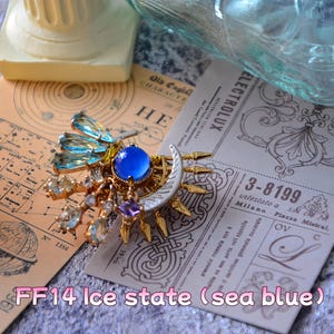 May include: A sea blue brooch featuring a central blue stone, clear and blue crystals, and a silver crescent moon. It rests on vintage paper with text and decorative elements.