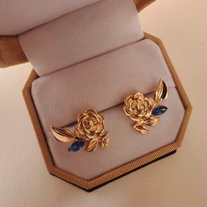 May include: A pair of gold-colored earrings shaped like roses with leaves and blue gemstone accents. The earrings are displayed inside a velvet-lined jewelry box with a gold-colored exterior.