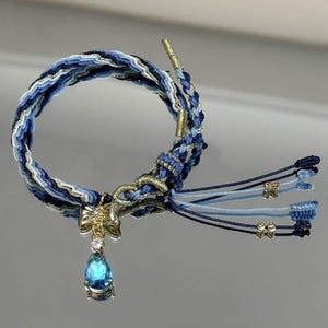 Può includere: Un braccialetto intrecciato blu e bianco con un ciondolo a forma di fiocco dorato e un pendente a goccia di pietra preziosa blu. Il braccialetto ha un design intrecciato con sfumature di blu e bianco e dettagli dorati.