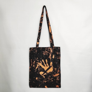 Può includere: Una borsa tote nera caratterizzata da un motivo candeggiato e maculato su tutto il tessuto. Al centro della borsa è presente una grafica prominente a forma di impronta di mano color arancione chiaro. Include due lunghi manici abbinati.