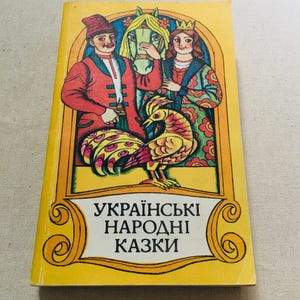 Pode incluir: Um livro retangular com capa amarela apresentando uma ilustração colorida de uma pessoa com casaco vermelho, um cavalo, uma pessoa com vestido verde estampado e coroa, e um pássaro. O texto "УКРАЇНСЬКІ НАРОДНІ КАЗКИ" está impresso em preto.