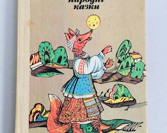Puede incluir: Una portada de libro vintage con una ilustración colorida de un zorro con un vestido tradicional ucraniano y sosteniendo un sol amarillo. El título del libro es "Семлитоцка Українські народні казки", que se traduce como "Cuentos populares de Ucrania".