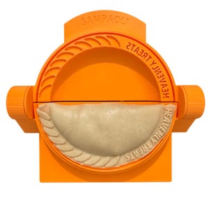 Puede incluir: Un molde para empanadillas naranja con el texto "SAMPAOLI" y "HEAVENLY TREATS". El molde está relleno con una empanadilla de color claro. El molde tiene un diseño circular con un borde ondulado.