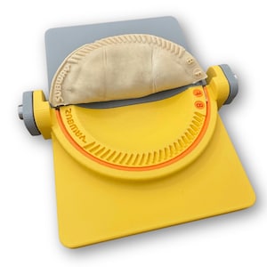 May include: A yellow and gray empanada maker with a semi-circular design. The maker has a handle on each side and the word "Subway" is printed on the top. An uncooked empanada is placed inside the maker.